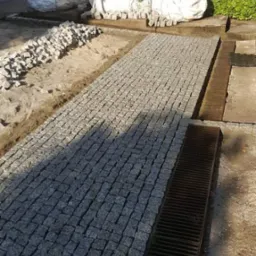 Fragment ścieżki z szarej kostki granitowej w trakcie układania, widoczne worki z materiałem i system odprowadzania wody.