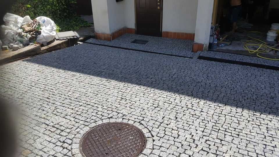 Szara kostka granitowa ułożona na podjeździe przed domem z widocznymi elementami odwodnienia liniowego i studzienką kanalizacyjną. W tle widoczne narzędzia i worki z materiałami budowlanymi.