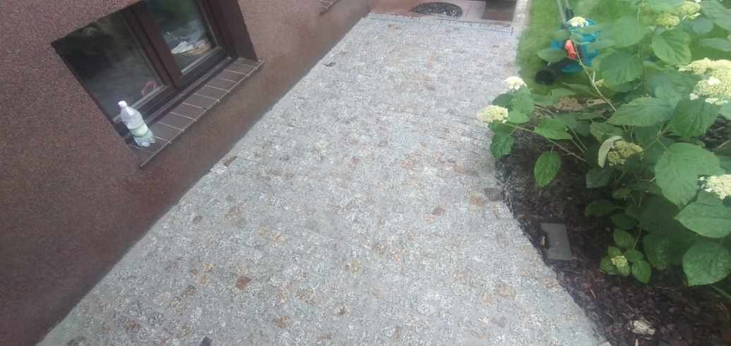 Ścieżka z jasnoszarej kostki granitowej prowadząca wzdłuż ściany domu z brązową elewacją, obok rabaty z kwitnącymi hortensjami i butelką wody na parapecie okna.