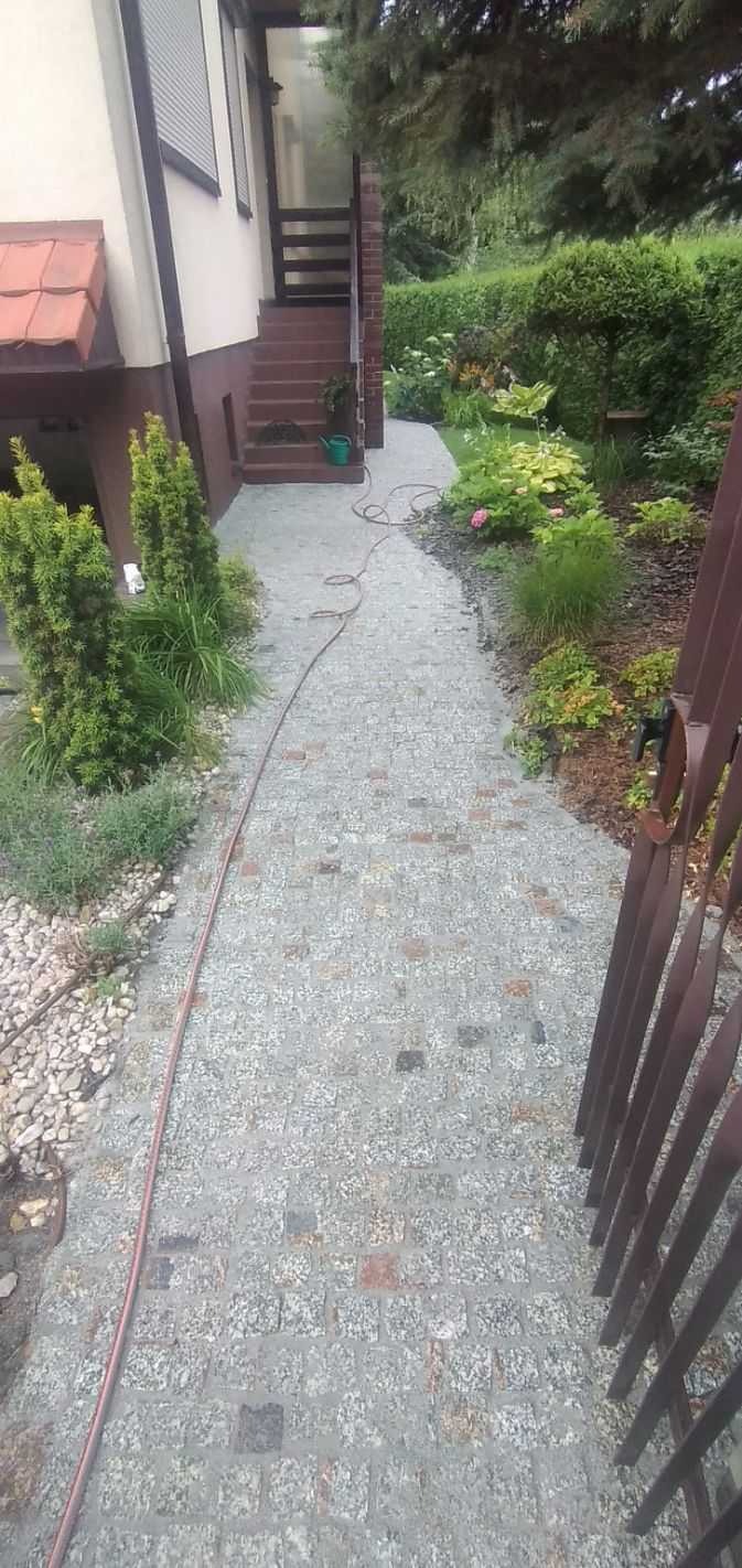 Ścieżka z kostki granitowej prowadząca od schodów domu do ogrodu, widoczny wąż ogrodowy i fragment metalowego ogrodzenia.