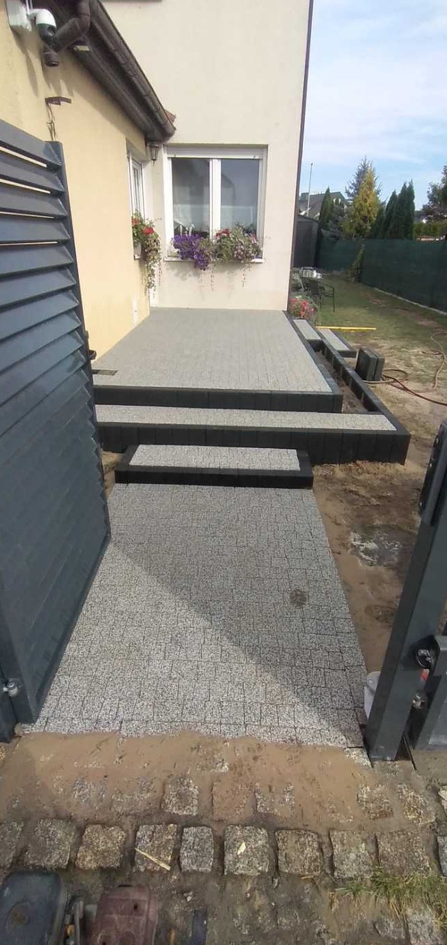 Nowo wykończony taras z szarej kostki granitowej i schodami z czarną obwódką, prowadzący od furtki do wejścia domu z oknem ozdobionym kwiatami.