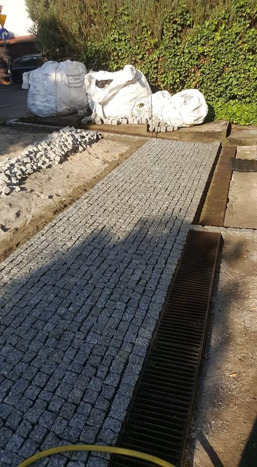 Fragment ścieżki z szarej kostki granitowej w trakcie układania, widoczne worki z materiałem i system odprowadzania wody.
