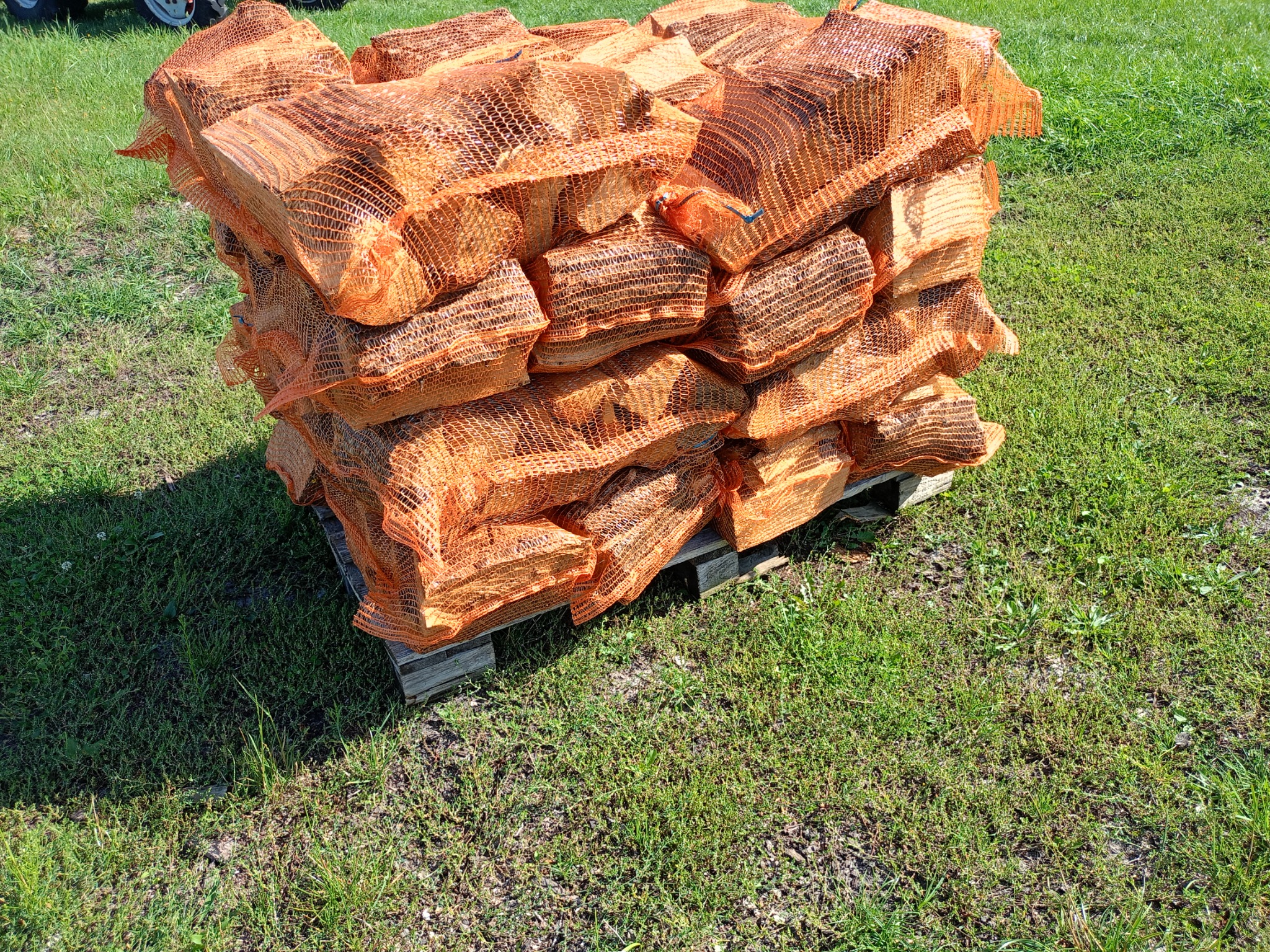 Stos worków z drewnem kominkowym ułożonych na drewnianej palecie na trawie, widoczne pomarańczowe siatki i fragment ciągnika w tle.