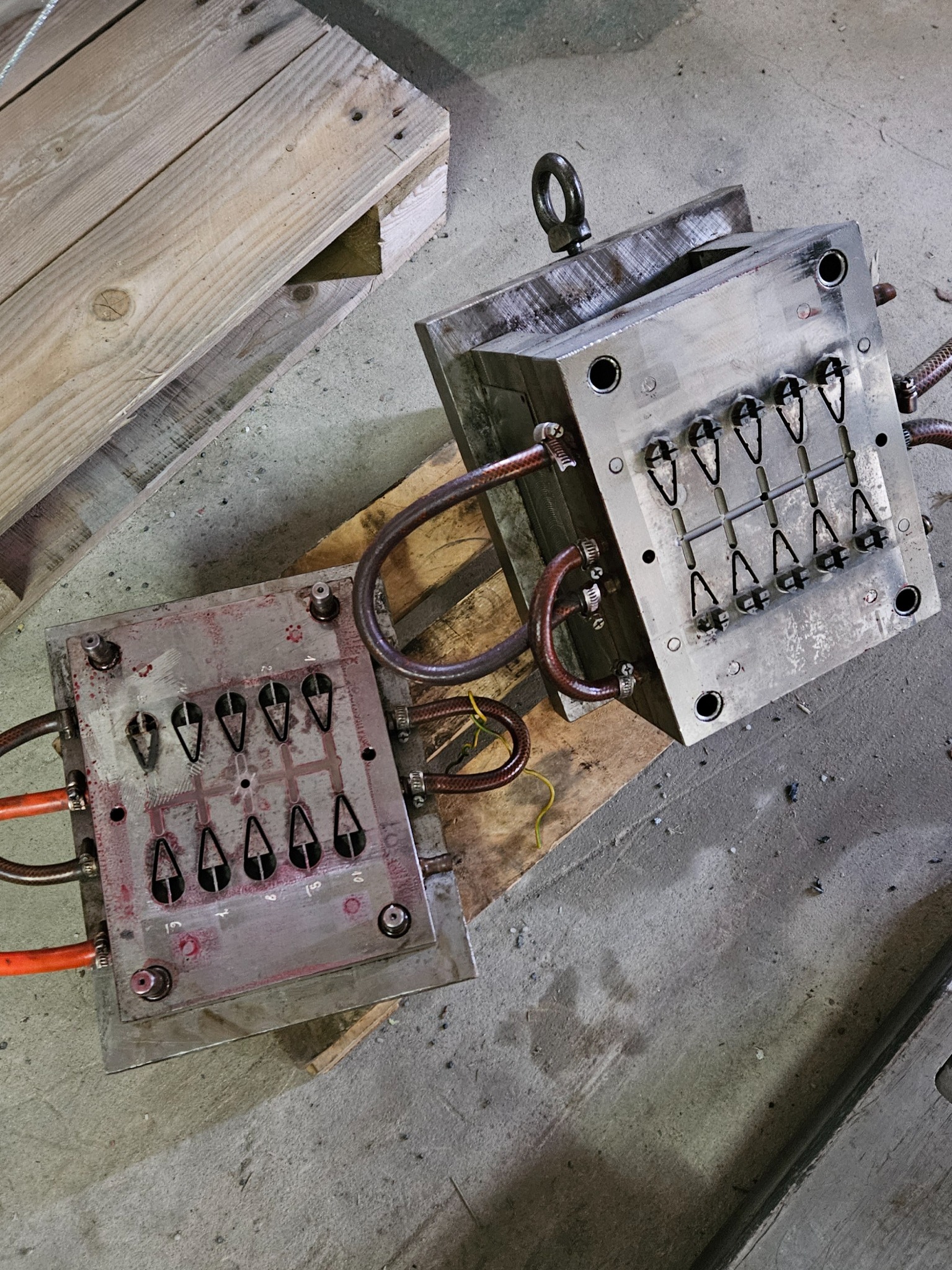 Dwie części metalowej formy wtryskowej z widocznymi kanałami chłodzącymi i otworami w kształcie strzałek, leżące na drewnianej palecie na betonowej podłodze.