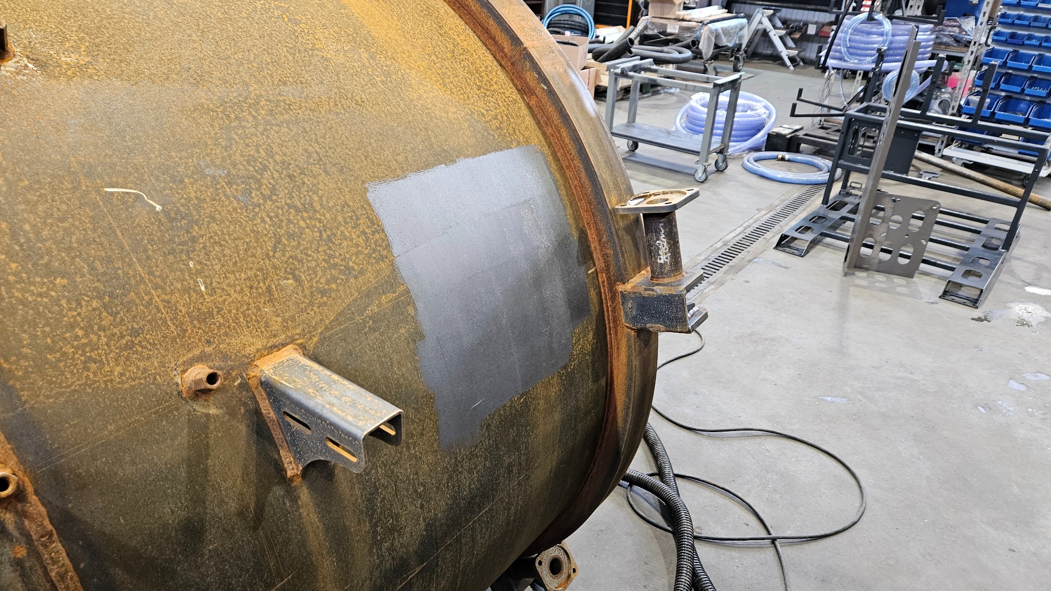 Fragment metalowego zbiornika z widoczną rdzawą powierzchnią i świeżo oczyszczonym, szarym prostokątnym obszarem, z metalowymi wspornikami, w tle warsztatowe otoczenie z wózkami i akcesoriami.