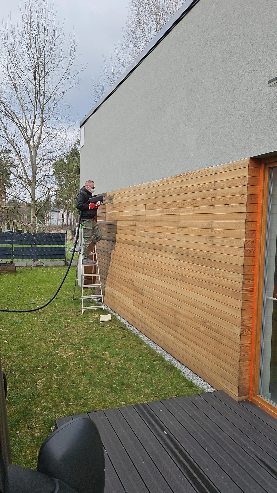 Pracownik na drabinie czyści drewnianą elewację domu za pomocą specjalistycznego sprzętu, widoczny fragment elewacji przed i po czyszczeniu.