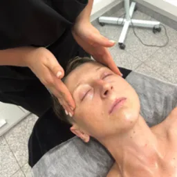 Kobieta z zamkniętymi oczami podczas masażu twarzy w salonie kosmetycznym, widoczne dłonie masażysty i fragment lampy kosmetycznej w tle.