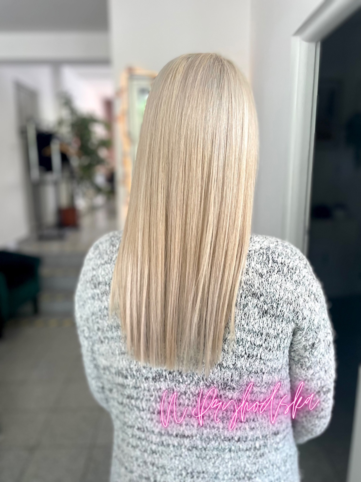 Tył głowy kobiety z długimi, prostymi, platynowymi blond włosami, noszącej szary, dziergany sweter, z rozmytym tłem wnętrza salonu.