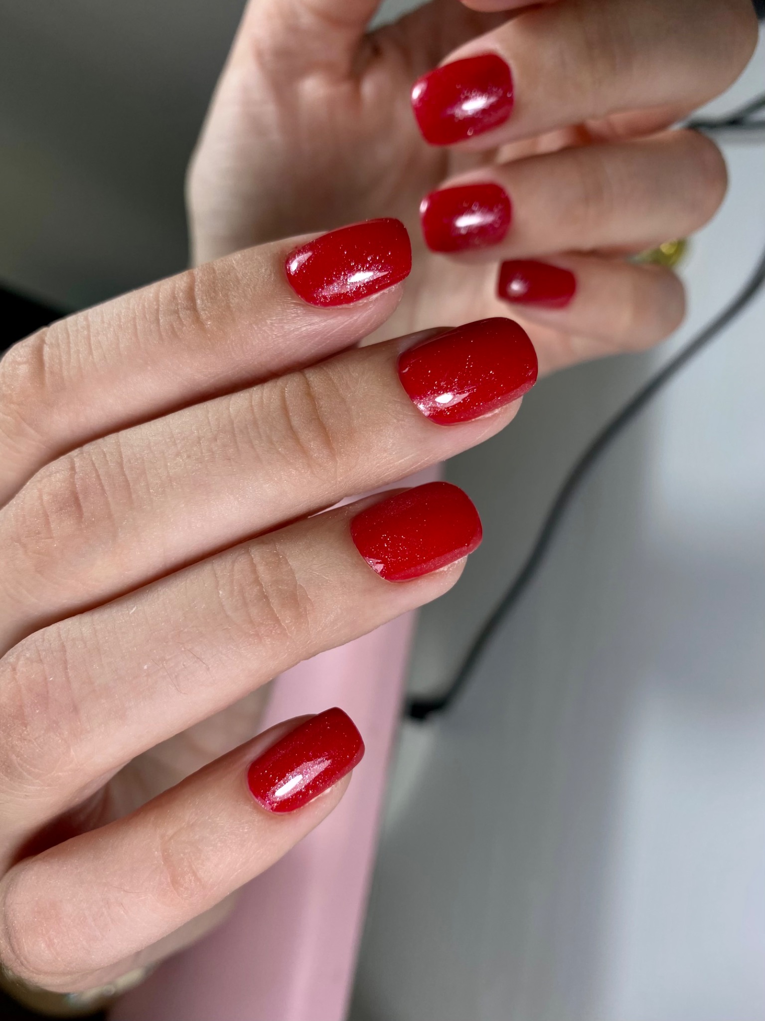 Dłonie z krótkimi, kwadratowymi paznokciami pomalowanymi na intensywny, błyszczący czerwony kolor z subtelnym brokatem, na tle jasnego blatu.