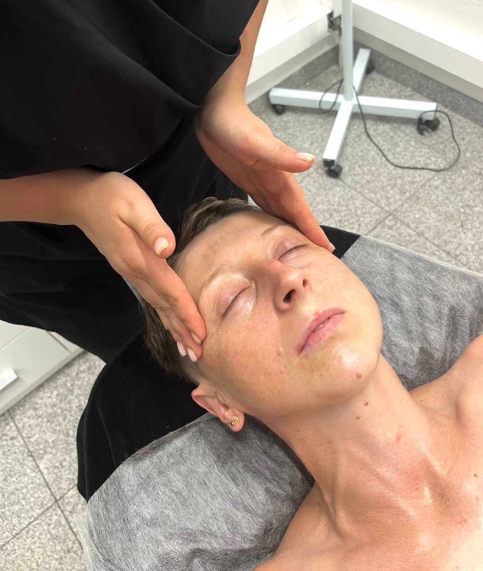 Kobieta z zamkniętymi oczami podczas masażu twarzy w salonie kosmetycznym, widoczne dłonie masażysty i fragment lampy kosmetycznej w tle.