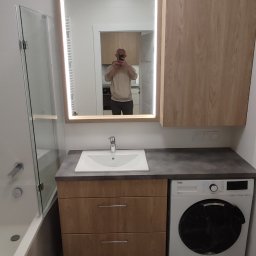 Zodczij - Nowoczesna łazienka z umywalką nablatową na szafce z dwiema szufladami, pralką Beko i lustrem z podświetleniem LED. W lustrze odbija się osoba robiąca zdjęcie na tle kuchni.