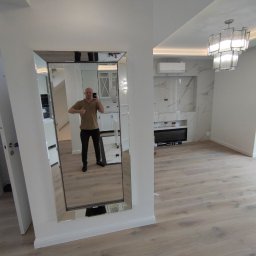 Zodczij - Duże lustro w ramie na ścianie w nowocześnie wykończonym wnętrzu z parkietem, widoczna osoba robiąca zdjęcie w lustrze oraz fragment kuchni w tle.