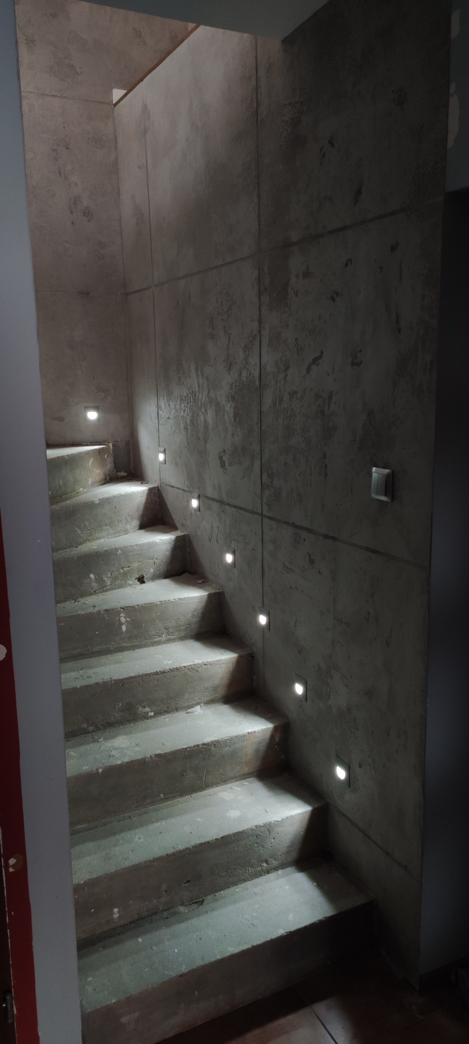 Betonowe schody z wbudowanym oświetleniem LED w ścianie, surowe wykończenie, widoczne łączenia płyt na ścianie, minimalistyczny design.