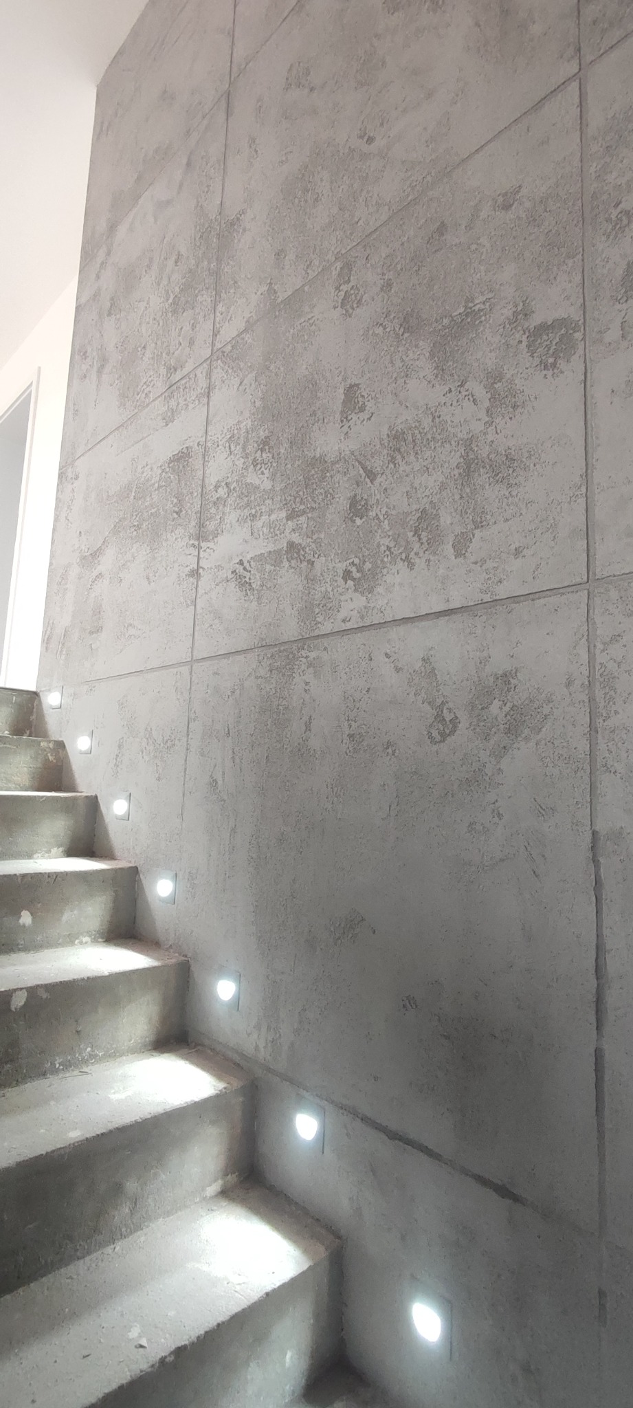 Ściana wykończona szarym tynkiem strukturalnym w stylu betonu architektonicznego w klatce schodowej w Warszawie, z oświetleniem LED wbudowanym w stopnie schodów.