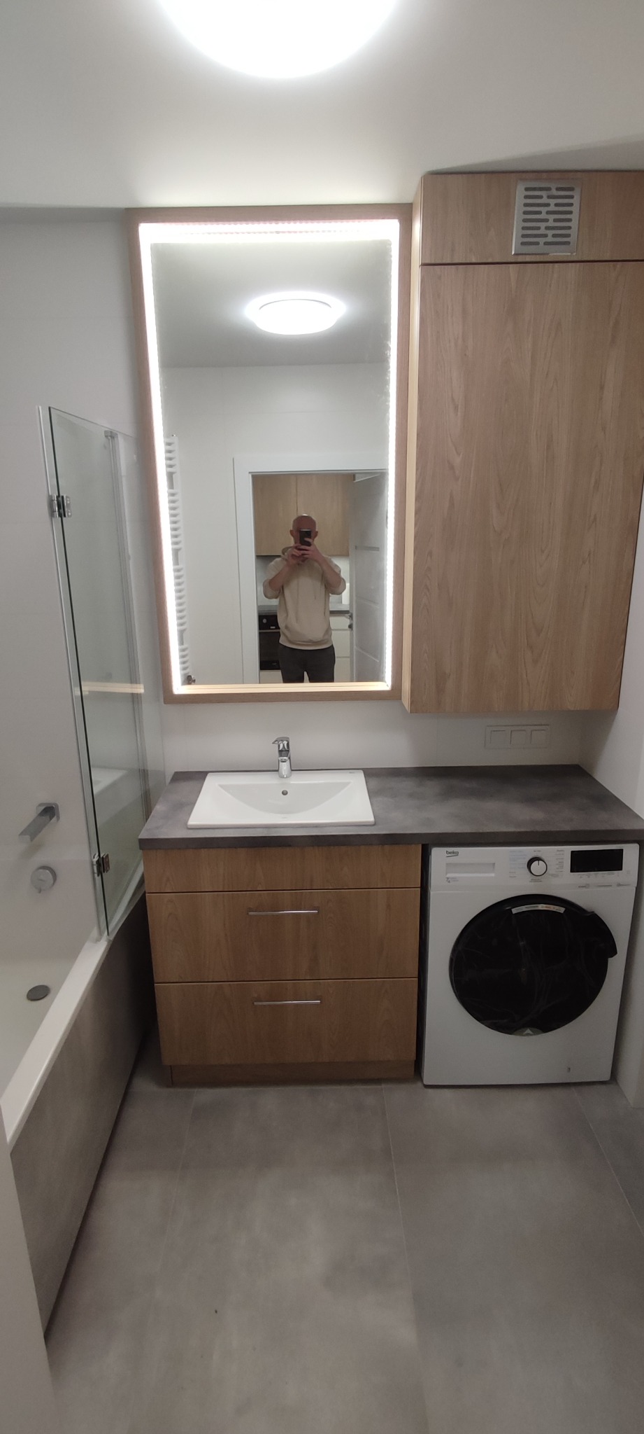 Nowoczesna łazienka z umywalką nablatową na szafce z dwiema szufladami, pralką Beko i lustrem z podświetleniem LED. W lustrze odbija się osoba robiąca zdjęcie na tle kuchni.