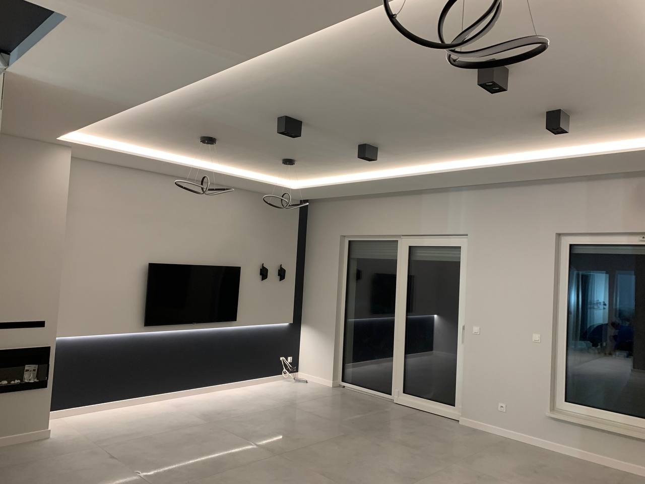 Nowoczesny salon w odcieniach szarości i bieli z podwieszanym sufitem z oświetleniem LED, minimalistycznym kominkiem i telewizorem na ścianie. Widoczne designerskie lampy sufitowe i duże okno...