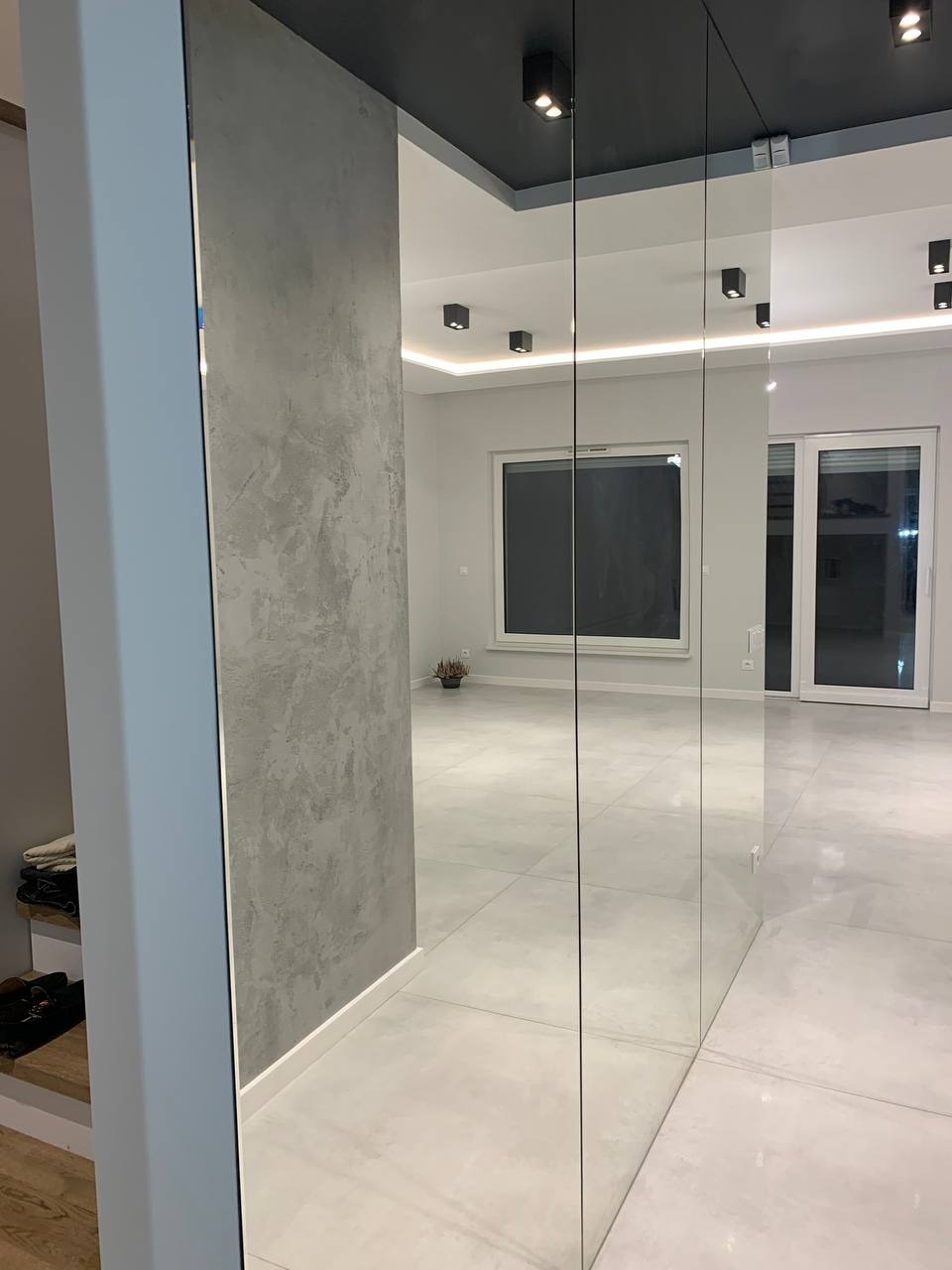 Nowoczesne wykończenie wnętrza: ściana z efektem betonu, lustrzane drzwi przesuwne i szara podłoga z płytek w przestronnym salonie z oknami.