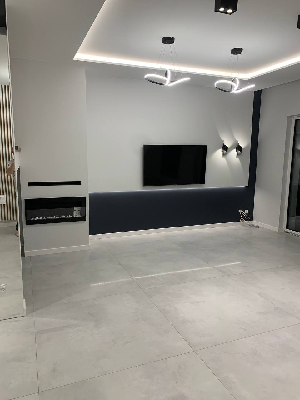 Nowoczesny salon z szarymi płytkami podłogowymi, podwieszanym sufitem z oświetleniem LED, czarnym telewizorem na ciemnogranatowej ścianie i minimalistycznym kominkiem.