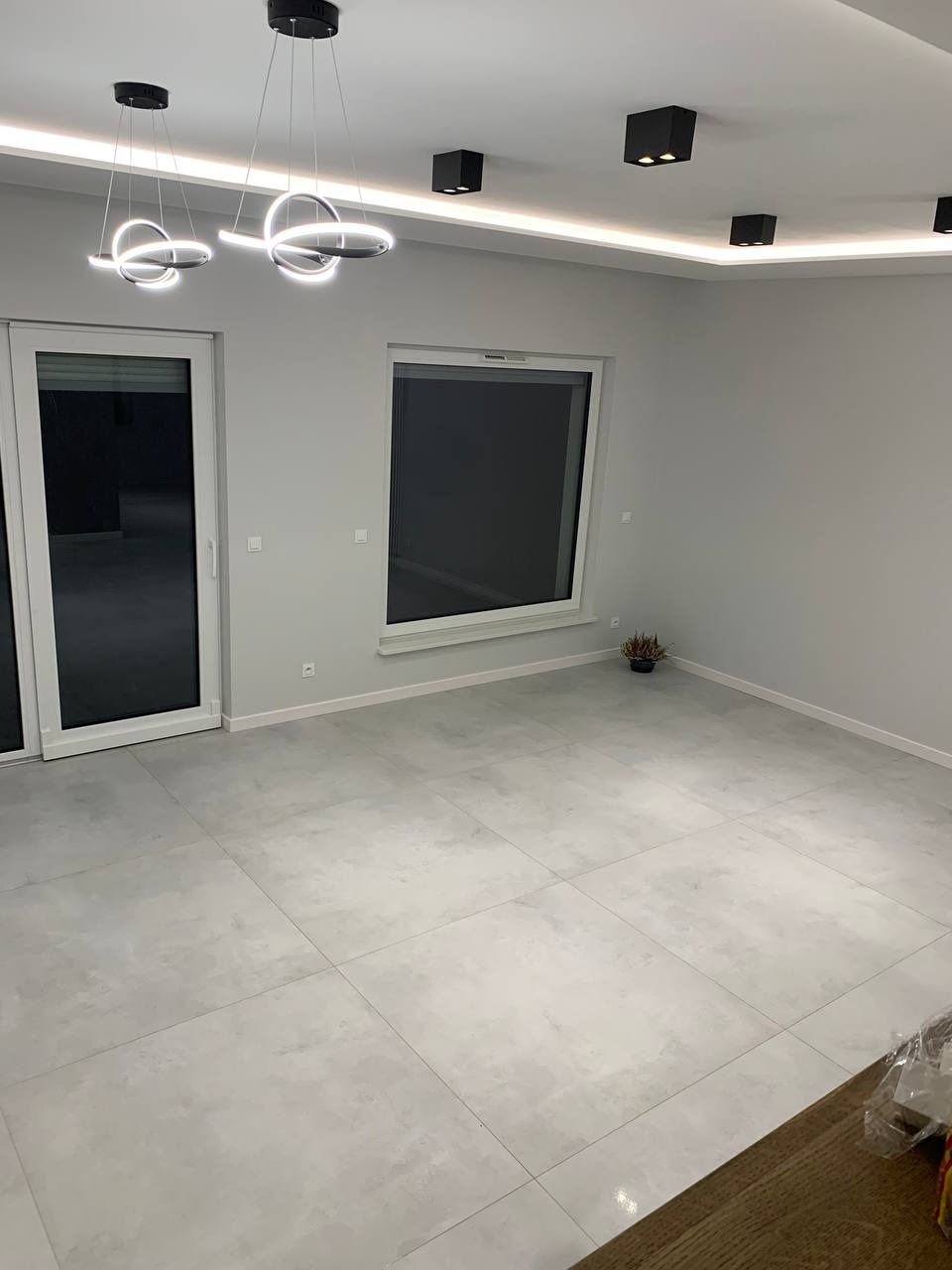 Jasne, przestronne wnętrze po remoncie z szarymi płytkami podłogowymi, nowoczesnym oświetleniem sufitowym, dużym oknem i rośliną doniczkową w rogu.