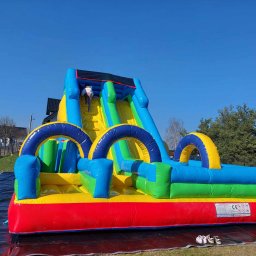 Easy Lingua - Kolorowa, dmuchana zjeżdżalnia z dzieckiem wspinającym się po schodach, rozłożona na czarnej plandece na trawie w słoneczny dzień.