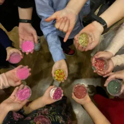 Grupa dzieci trzymająca w dłoniach przezroczyste pojemniki z kolorowymi masami plastycznymi i brokatem, widok z góry.