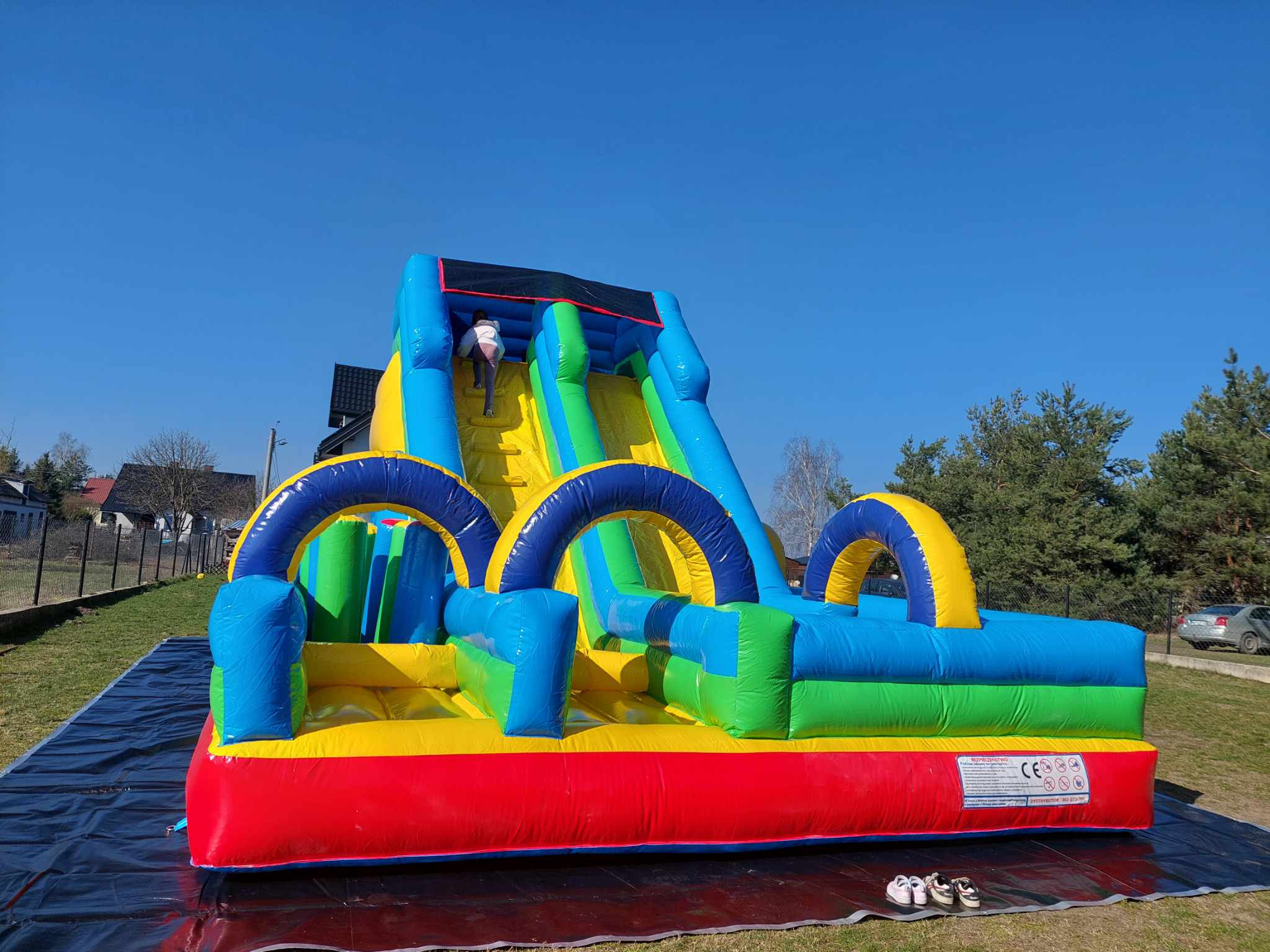 Kolorowa, dmuchana zjeżdżalnia z dzieckiem wspinającym się po schodach, rozłożona na czarnej plandece na trawie w słoneczny dzień.