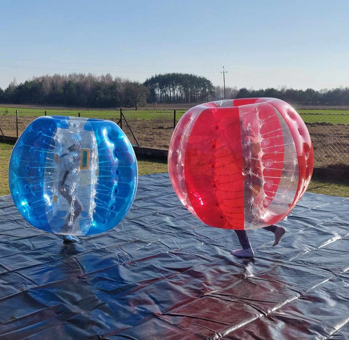 Dwie osoby wewnątrz przezroczystych kul sferycznych: jedna niebieska, druga czerwona, stojące na czarnej, błyszczącej macie na trawiastym terenie z drzewami w tle.