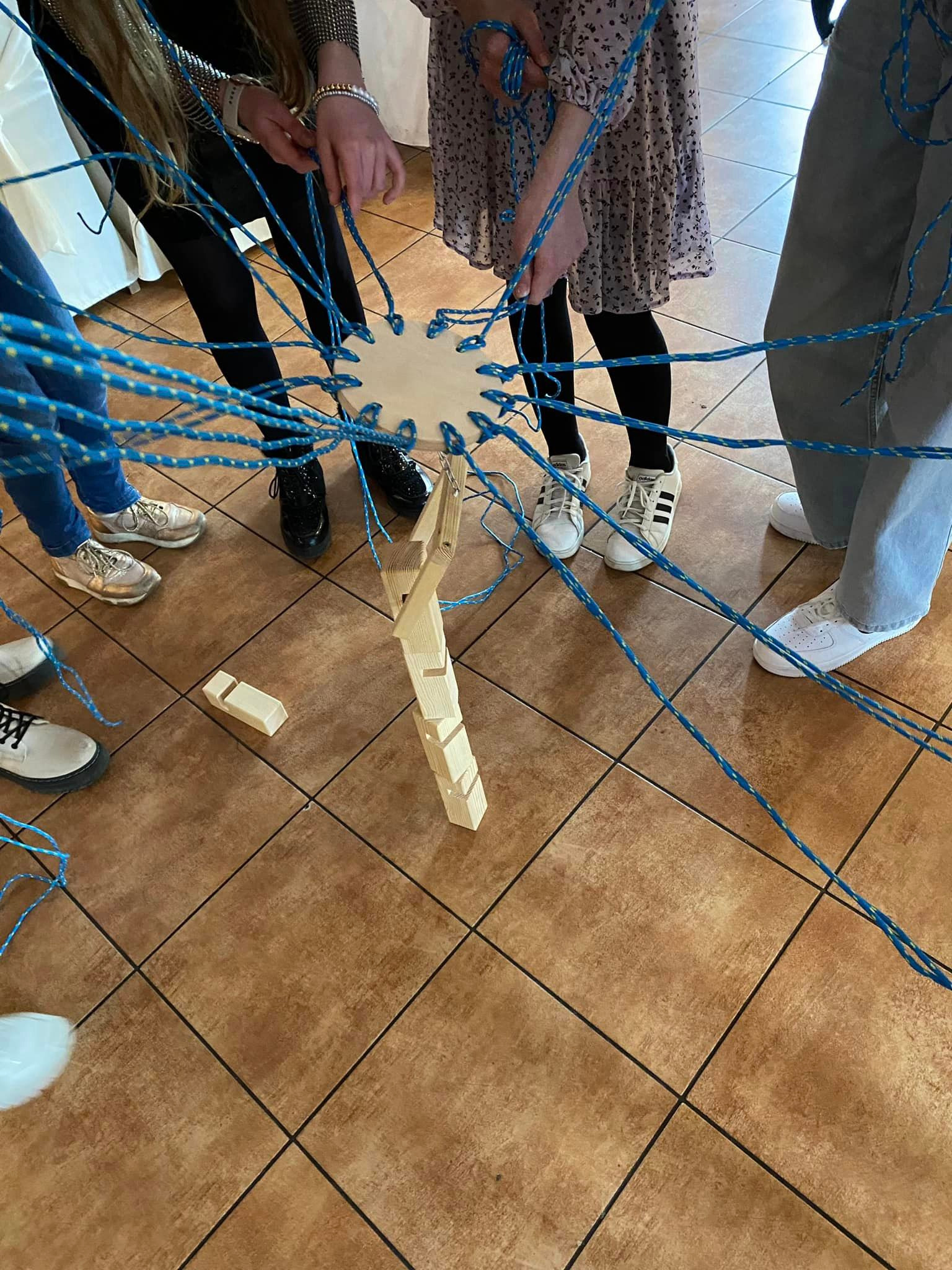 Grupa dzieci i dorosłych współpracuje przy grze zespołowej z drewnianą konstrukcją i niebieskimi sznurkami, skupieni na zadaniu.
