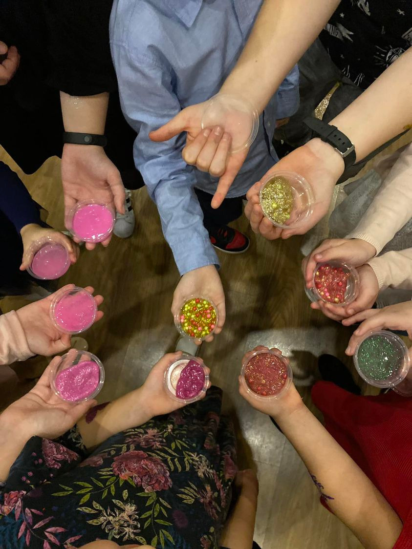 Grupa dzieci trzymająca w dłoniach przezroczyste pojemniki z kolorowymi masami plastycznymi i brokatem, widok z góry.