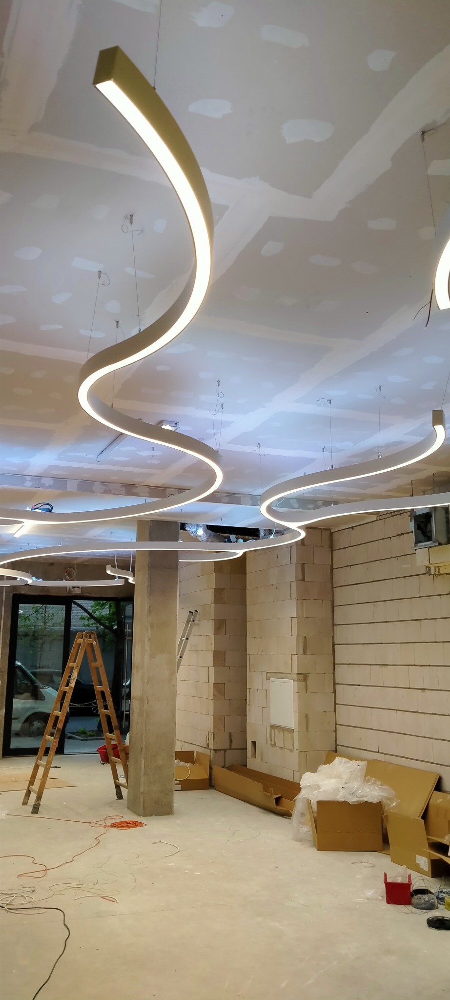 Faliste, podwieszane oprawy oświetleniowe LED w surowym wnętrzu. Widoczne drabiny i materiały budowlane wskazują na trwający montaż.