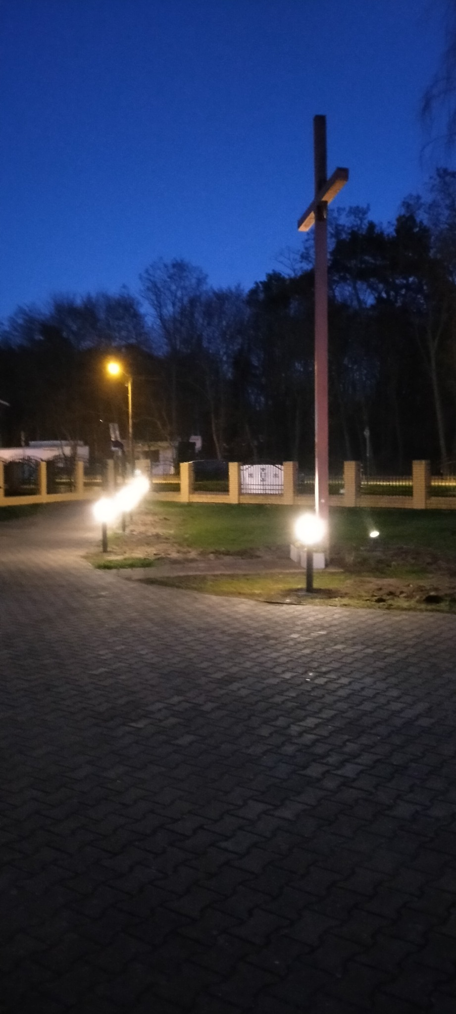 Świecące kule oświetleniowe wzdłuż brukowanej ścieżki prowadzącej do ogrodzenia z krzyżem w tle, oświetlonym dodatkową lampą uliczną, sfotografowane o zmierzchu.