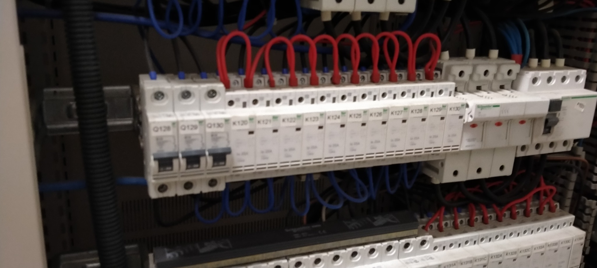 Rozdzielnica elektryczna z zainstalowanymi bezpiecznikami automatycznymi i okablowaniem w kolorach czerwonym i niebieskim, widoczne oznaczenia na obudowach.