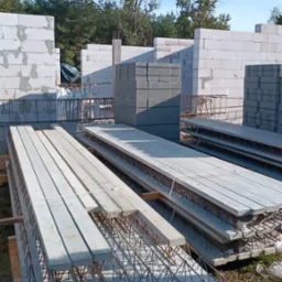 Konstrukcja budynku w trakcie budowy z betonu komórkowego, widoczne prefabrykowane belki stropowe ze zbrojeniem, ułożone na ziemi w oczekiwaniu na montaż.