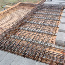 Siatka zbrojeniowa ułożona na deskowaniu, widok z góry, z bloczkami betonowymi na obrzeżach, w tle częściowo widoczny budynek z niebieskim dachem.