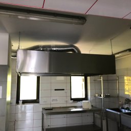JD Elektroinstalacje - Wnętrze kuchni przemysłowej z okapem wentylacyjnym zawieszonym na łańcuchach, widoczne gniazda siłowe i tabliczka informacyjna 'Obróbka Termiczna'.