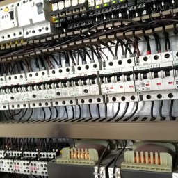 JD ELEKTORINSTALACJE JAROSŁAW DZIADYNAK - Szczegółowe ujęcie wnętrza szafy sterowniczej z licznymi przekaźnikami, bezpiecznikami i transformatorami, widoczne przewody i oznaczenia na elementach.