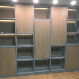 JD ELEKTORINSTALACJE JAROSŁAW DZIADYNAK - Wielofunkcyjny regał z szarymi ramami i drewnopodobnymi panelami, podświetlony od góry ciepłym światłem LED, stojący na drewnianej podłodze.