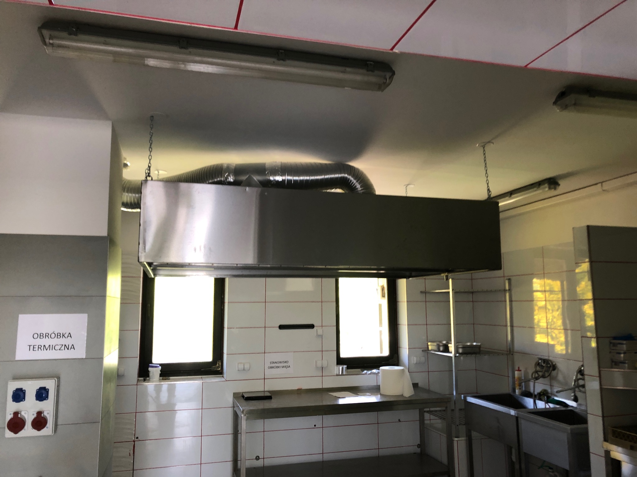 Wnętrze kuchni przemysłowej z okapem wentylacyjnym zawieszonym na łańcuchach, widoczne gniazda siłowe i tabliczka informacyjna 'Obróbka Termiczna'.