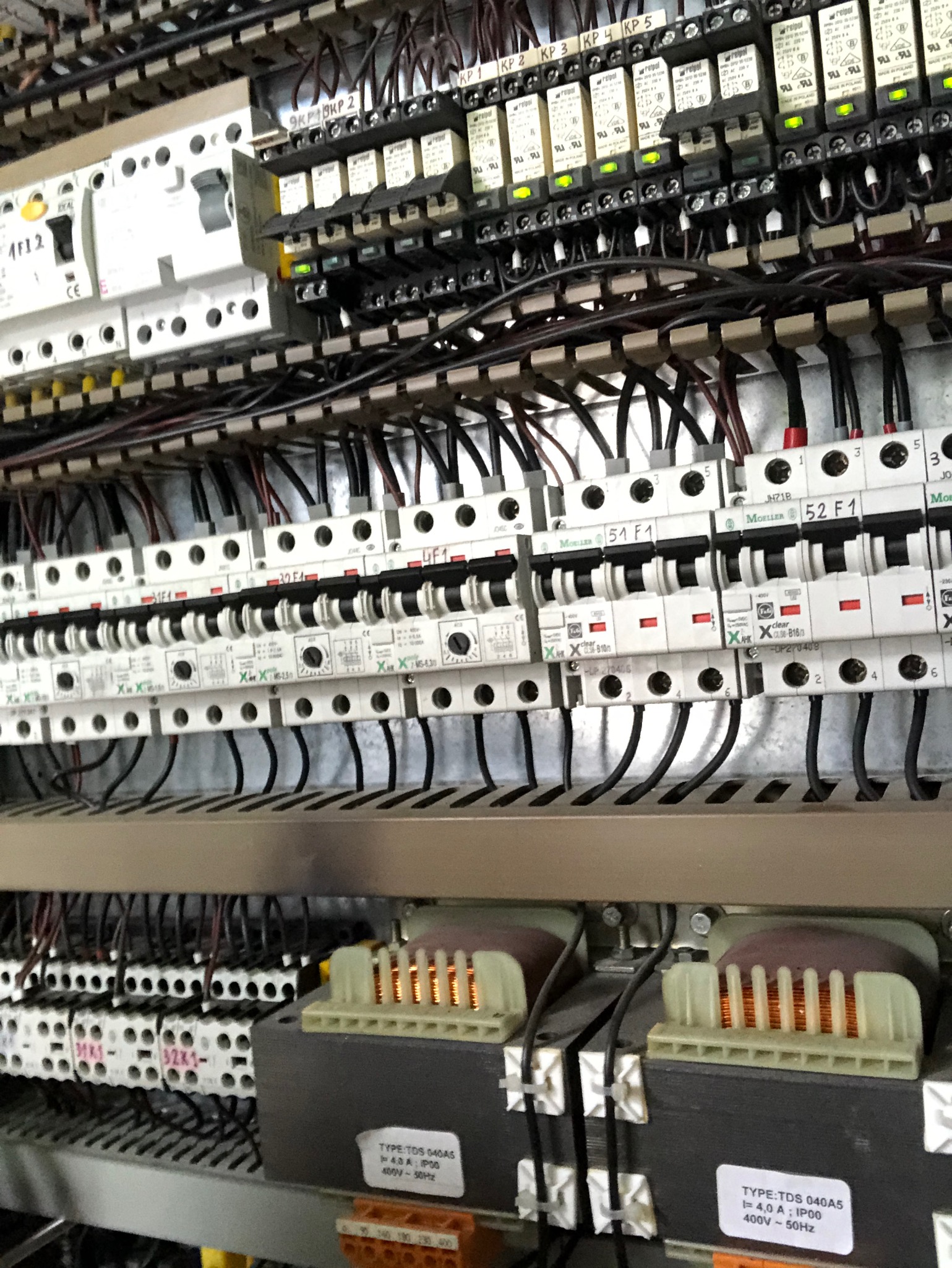 Szczegółowe ujęcie wnętrza szafy sterowniczej z licznymi przekaźnikami, bezpiecznikami i transformatorami, widoczne przewody i oznaczenia na elementach.