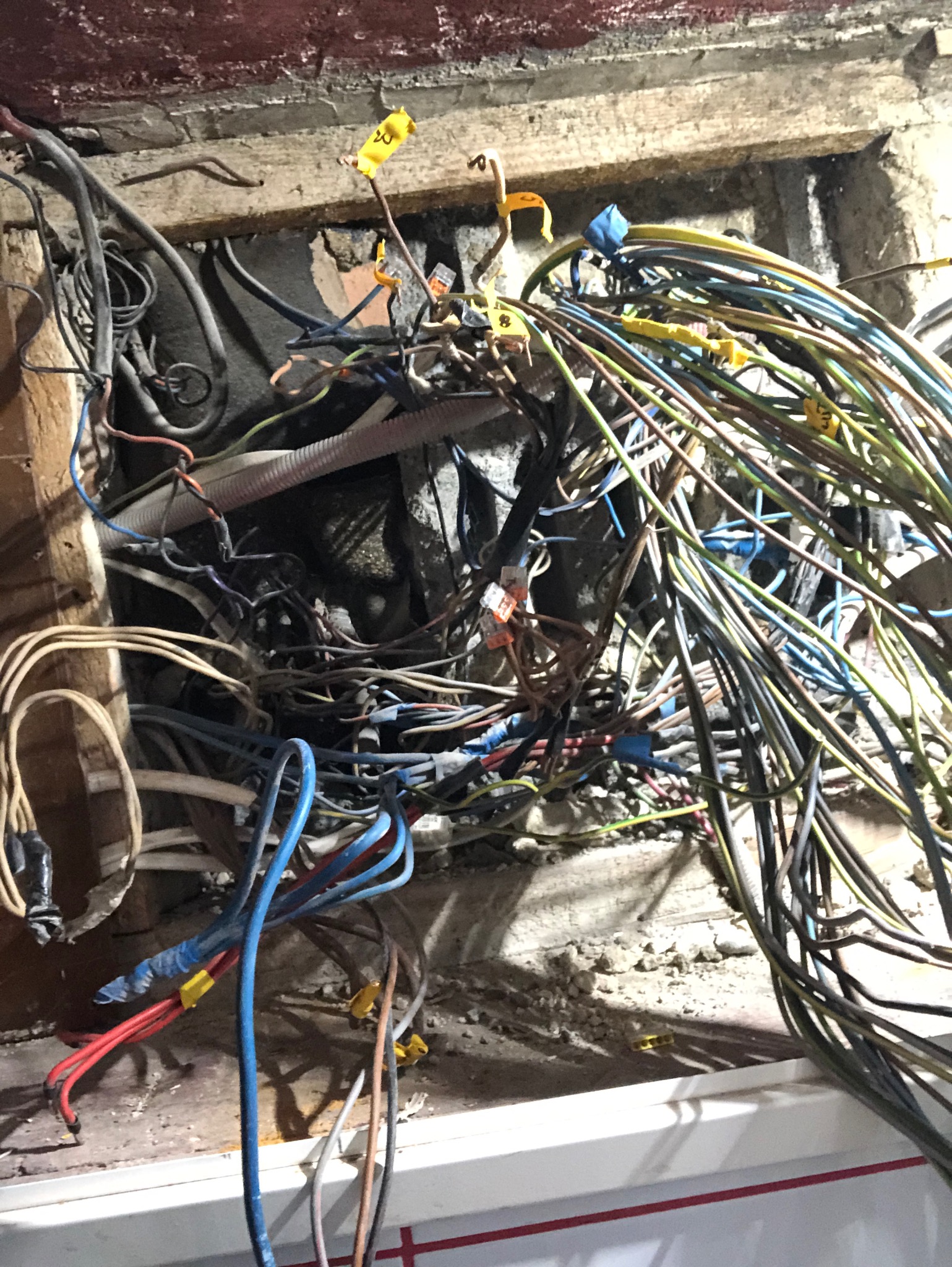 Skomplikowany węzeł różnokolorowych przewodów elektrycznych w ścianie, z widocznymi złączkami WAGO i żółtymi oznaczeniami, w surowym, niedokończonym otoczeniu.