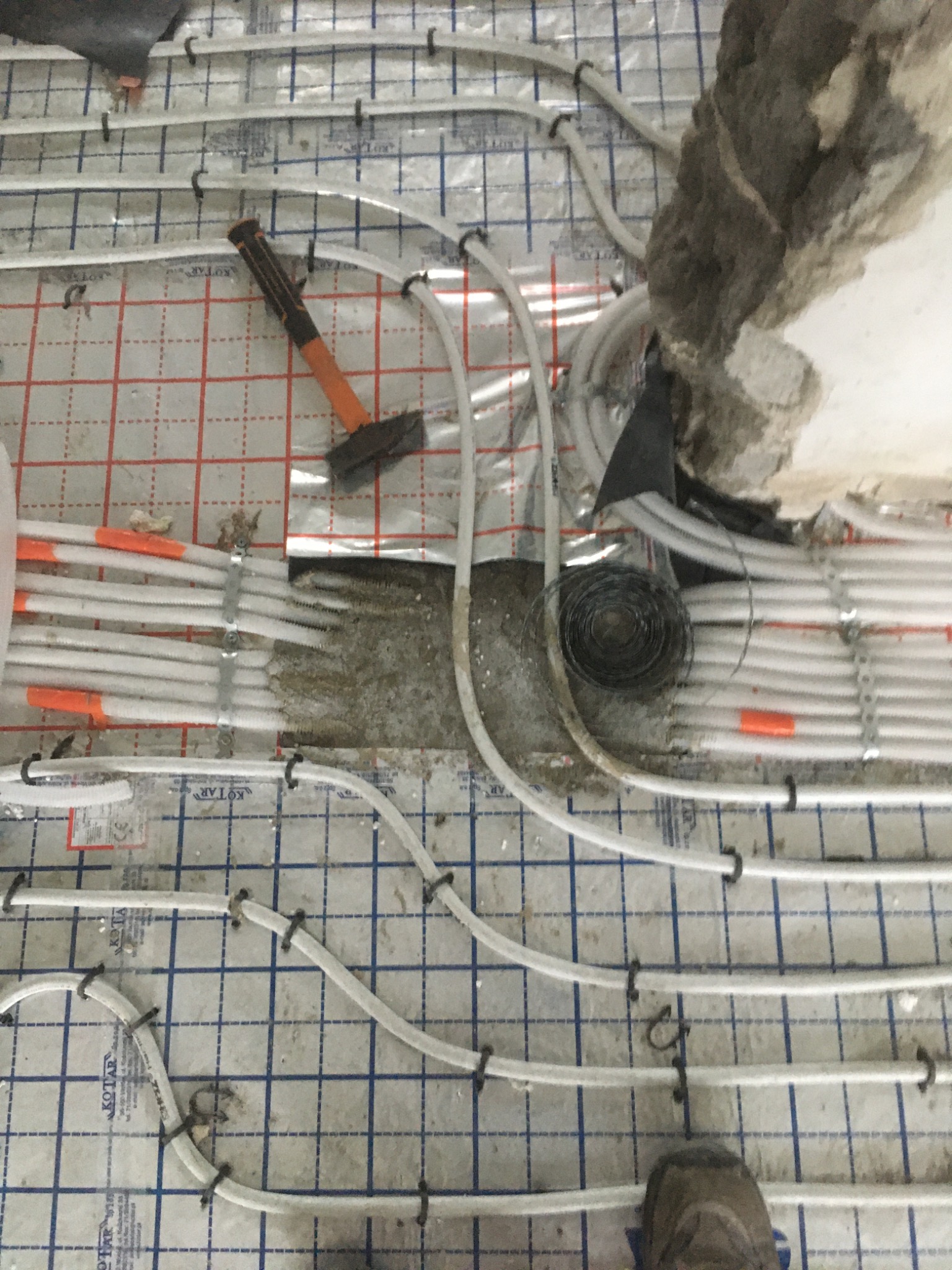 Montaż ogrzewania podłogowego: białe rury mocowane do folii aluminiowej z siatką, widoczny młotek z pomarańczowym trzonkiem i zwój drutu, fragment zdemontowanej ściany w tle, ujęcie z góry.