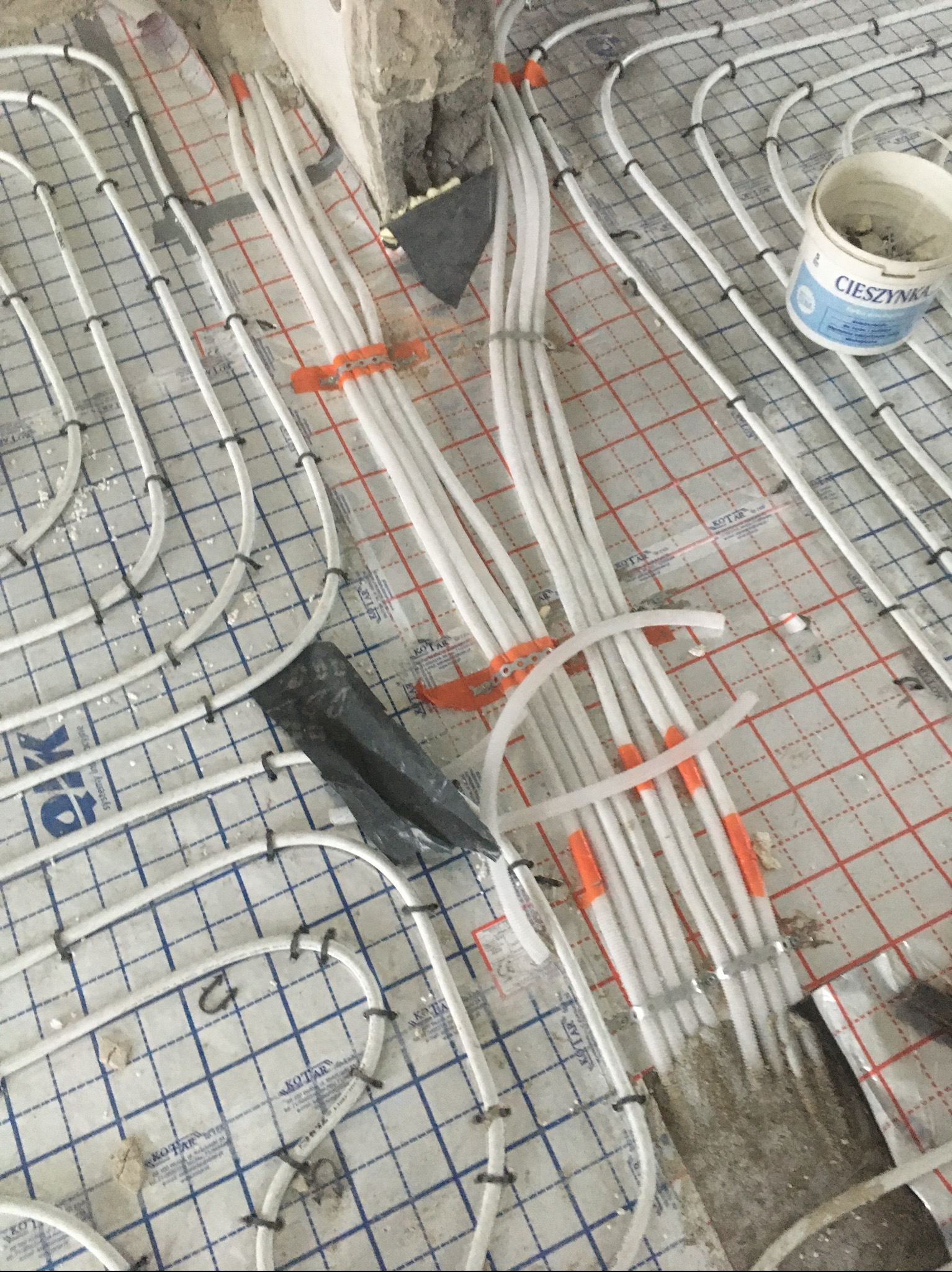 Białe rurki ogrzewania podłogowego przymocowane do folii z siatką, widoczny fragment ściany i wiadro z napisem Cieszynka.