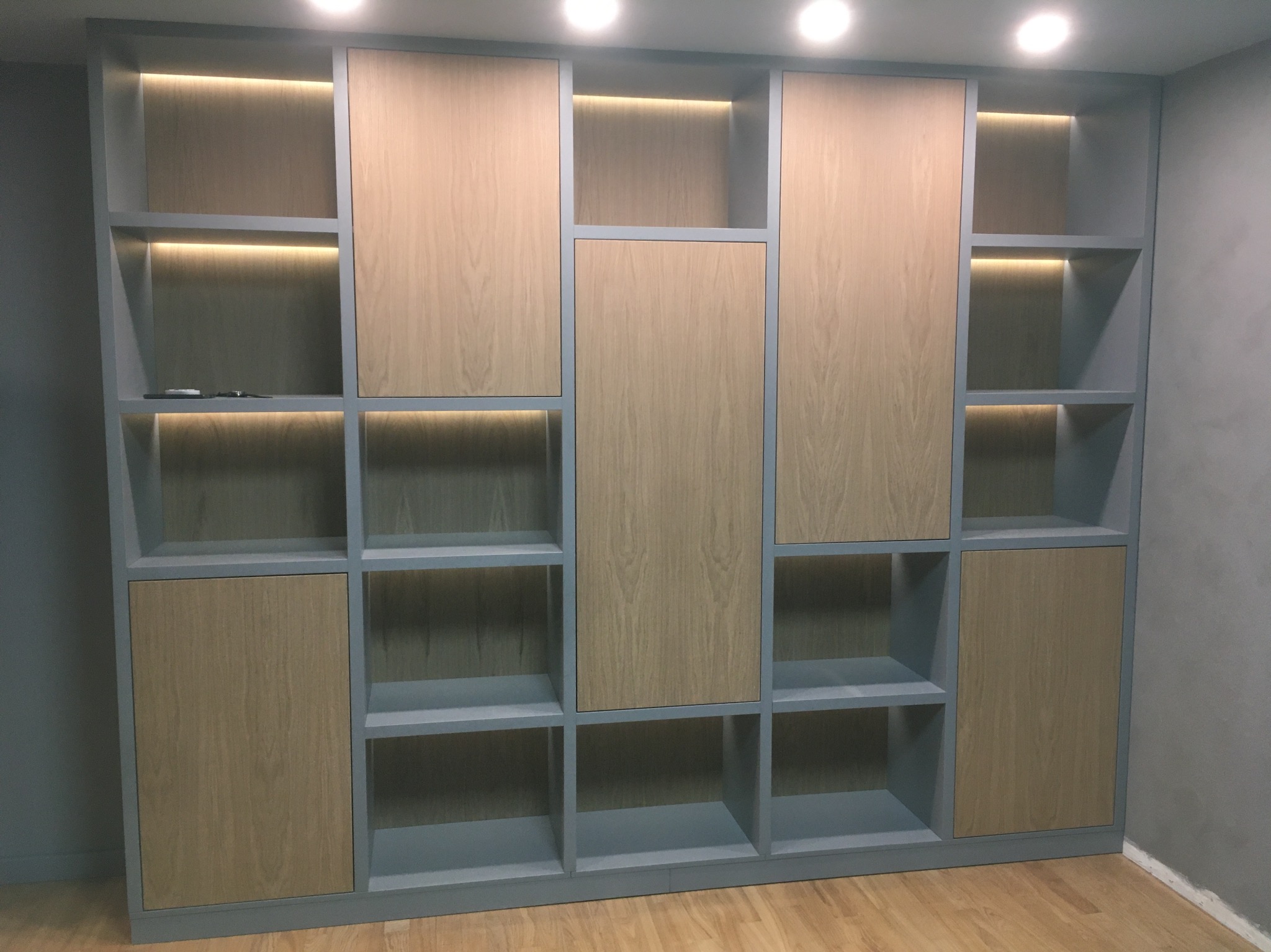 Wielofunkcyjny regał z szarymi ramami i drewnopodobnymi panelami, podświetlony od góry ciepłym światłem LED, stojący na drewnianej podłodze.