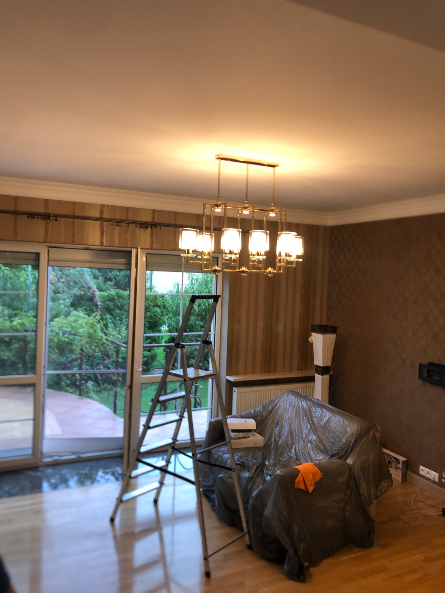 Nowoczesny żyrandol z kwadratowymi kloszami w salonie podczas montażu, drabina oparta o okno balkonowe, meble zabezpieczone folią malarską.