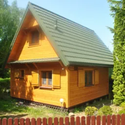 Niewielki, parterowy domek z drewna o żółtej elewacji i zielonym dachem, otoczony zielenią, widoczny zza niskiego, czerwonego płotku.