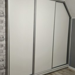 Mateusz Jakubczak - Trzydrzwiowa szafa przesuwna z białymi frontami i srebrnymi obramowaniami, wkomponowana we wnękę pod skosem sufitu, z widoczną ścianą z imitacji cegły.