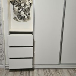 Mateusz Jakubczak - Biała szafa przesuwna z otwartą wnęką z trzema szufladami i koszulą w tropikalny wzór wiszącą na wieszaku, widoczna faktura drewnopodobnej podłogi.