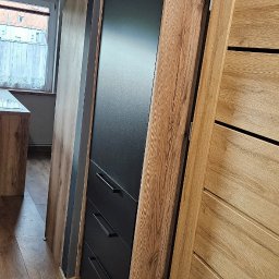 Mateusz Jakubczak - Wnętrze z zabudową: szafa z czarnymi frontami i trzema szufladami, obudowana drewnopodobną płytą, widoczna podłoga z paneli i fragment drzwi z drewnianą okleiną.