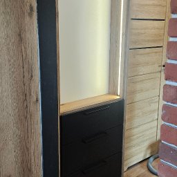 Mateusz Jakubczak - Wnęka z szafką na buty i wieszakami, wykończona w drewnie z trzema czarnymi szufladami i oświetleniem LED, widok z boku na korytarz z drewnianą podłogą i ceglaną ścianą.