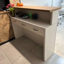 Mateusz Jakubczak - Biała lada recepcyjna z drewnianym blatem w salonie w Wałbrzychu, na blacie wazon z tulipanami i dekoracje, w tle sprzęt kosmetyczny.